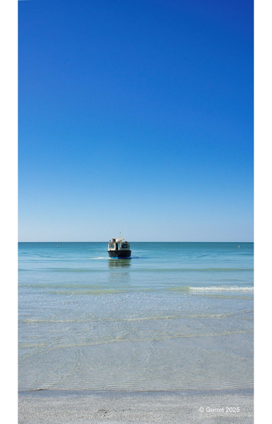 A lone boat approaches the shore under a clear blue sky, gently rippling the calm, turquoise ocean. The sandy beach is serene and inviting.