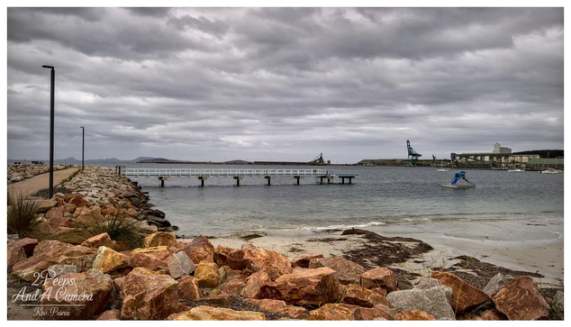 A wide angle landscape photograph of the Esperance Tanker Jetty area on a cloudy, overcast day.

In the foreground, a pile of large, reddish-orange granite boulders forms a natural barrier leading up to a paved walking path on the left.

The water is calm, meeting a small sandy beach patch with seaweed. A long, low white jetty extends into the water toward the center.

In the background, industrial elements of the working port are visible, including a large green crane and port buildings, backed by distant hills under a vast, gray sky.