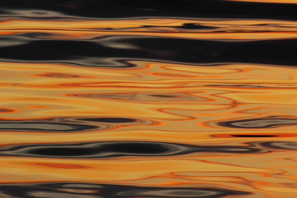 Photograph of sunset light reflections on the water with horizontal swirls and patterns of yellow, orange, grey, and black.