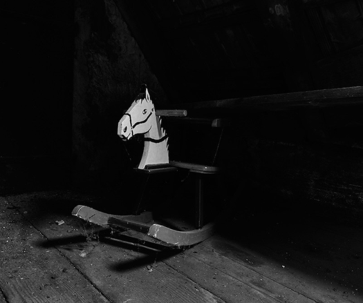 Eine dunkle Szene auf einem alten Dachboden. Ein altes Schaukelpferd das unter einer Ablage hervor scheint. Es sind nur der vordere Teil mit dem Kopf zu sehen, der Rest verschwindet in der Dunkelheit. Das Foto ist in Schwarzweiß.