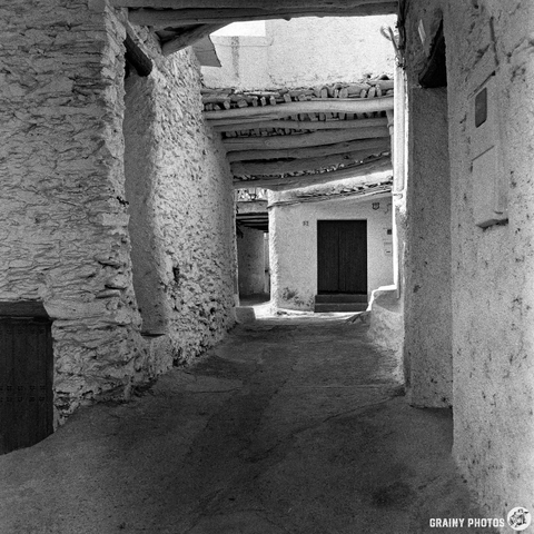 A narrow, winding alleyway in Capilerilla, Spain, with white stone walls and a rustic wooden tinao, leads to a closed wooden door; shadows and light create a peaceful, timeless atmosphere in this black and white photo.