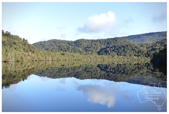 Colour landscape photograph of the Gordon River in Tasmania, signed by Kev Peirce Photography.  The image features a wide expanse of still, dark water perfectly reflecting the dense, deep green temperate rainforest covering the steep hillsides that frame the river.  The reflection is almost a perfect mirror image, including the small patches of white cumulus clouds in the pale blue sky above. The scene captures the tranquility and pristine nature of the remote wilderness.