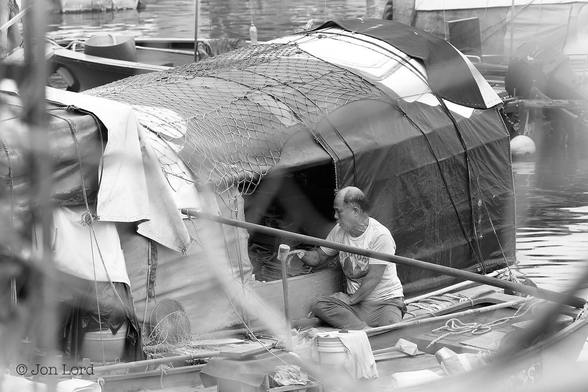 This is a black and white street photo in landscape format of a Chinese man sitting in a dinghy and looking in on a small houseboat. Causeway Bay, Hong Kong (2013).

To the lower right of centre is our Chinese man, nearly bald, wearing a white t-shirt and loose, long trousers. He is leaning in a rectangular opening of a home-built houseboat. The houseboat is nearly at right angles to the camera and while it is not all in view its probably about six metres long. The boats covering is made up of numerous awnings and plastic sheets slung over various metal frames and secured to the boat with a number of ropes and nets. Our Chinese man has rowed over to the houseboat to check up and talk to an elderly man (out of sight) living on board. Surrounding this scene are numerous other houseboats making up this floating village in the Causeway Bay Typhoon Shelter, Hong Kong.

