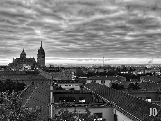 Fotografía en blanco y negro de un paisaje urbano al atardecer o amanecer, vista desde una perspectiva elevada. En el fondo, a la izquierda, domina la silueta oscura de una gran catedral con una cúpula prominente y una torre alta y elaborada. El cielo está cubierto por una densa capa de nubes onduladas y texturizadas. En el primer plano y el medio, se aprecian numerosos tejados oscuros de edificios urbanos, con algunas ventanas iluminadas. En el horizonte distante, se extiende el resto de la ciudad con luces tenues y una atmósfera brumosa.