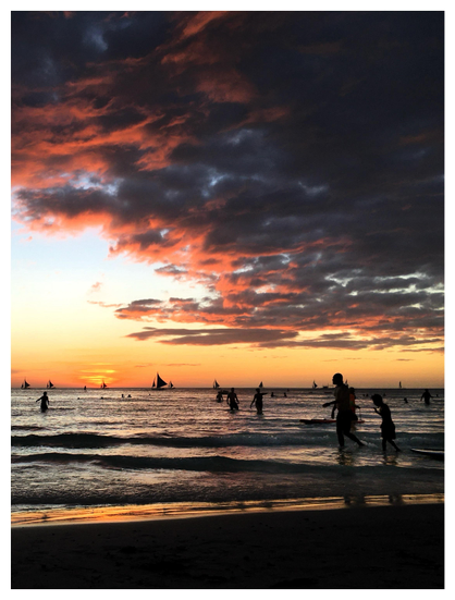 Photo of an island beach at sunset. Waves gently lap against the dark sand. People stroll in the ankle-deep water, silhouetted in the golden light. Sailboats line the horizon. The sun, partially behind cloud streaks, casts a warm glow over the sea and paints a luminous orange lining on a shadowy mass of low clouds.