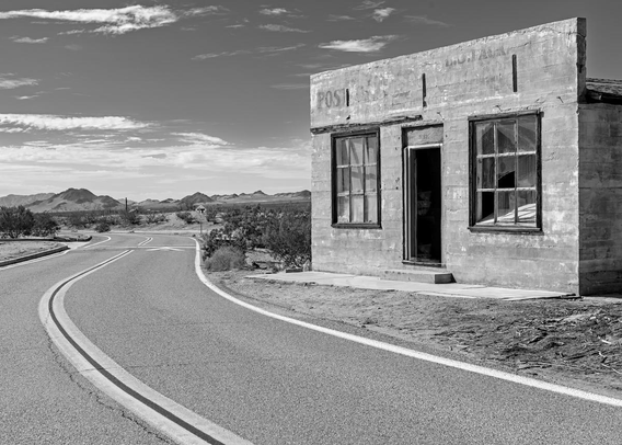 A black and white landscape photo of an old abandoned building on a deserted desert highway. The building is small and was apparently a Post Office given that the word "Post" is seen on the far left corner of the building. There's a central doorway (no door) and large windows on either side of the door. Most of the glass is still in the windows. The two lane road, with a double stripe down the center winds around from the bottom of the frame to the center and then off to the left. The sky has clouds on the horizon above some distant hills.