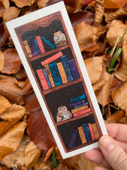 bookmark of a very whimsical dark academia looking bookshelf. The shelves are lined with vsrious antiquarian books, and on the top shelf is a sweet little terrarium, and on the third shelf down is perched a studious little ruru owl drinking a cup of tea. Very witchy, very whimsy, extremely magical.