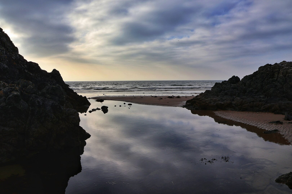 A serene coastal scene captured at what appears to be either dawn or dusk. The image showcases a tranquil, shallow tidal pool reflecting the soft hues of the sky, framed by rugged, dark rock formations on either side. Beyond the pool, a sandy beach stretches out towards the calm sea, where gentle waves lap the shoreline. The sky above is adorned with wispy, layered clouds, adding depth and texture to the scene.