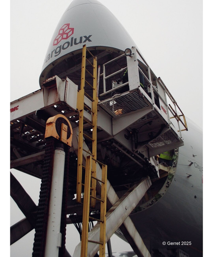 The image shows the nose of a cargo plane open with a loading platform and ladder in the foreground. The sky is overcast, creating a somber tone.
