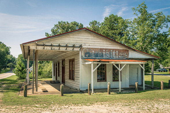 A weathered white wooden country store with horizontal lap siding and a red metal gabled roof. The single-story building features a covered front porch supported by wooden posts, with a wooden deck floor. A faded vintage Coca-Cola sign is visible on the facade above a rusty corrugated metal awning that extends over part of the porch. The structure has dark-framed windows and doors, including what appears to be a multi-paned storefront window. Wooden posts mark the edge of the porch area. The building sits on a grassy lot with mature green trees in the background under a partly cloudy blue sky. A gravel or dirt area is visible in the foreground, and a road can be seen to the left side of the property.