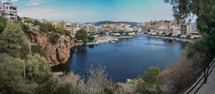 A scenic view of a harbor surrounded by buildings and rocky cliffs, with calm water reflecting the sky. There are boats moored along the shoreline, and lush greenery in the foreground.
