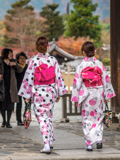 Dos jóvenes de espaldas caminando hacia los jardines del templo. Van vestidas con un kimono sencillo (yukata, creo que se llama) de color blanco y detalles rosados. Ambas llevan un obi (la faja del kimono) de color rosa y bolsos muy parecidos. El pelo con un moño bajo y algún adorno (un coletero, probablemente) con motivos florales. Ambas con calcetines blancos y sandalias.