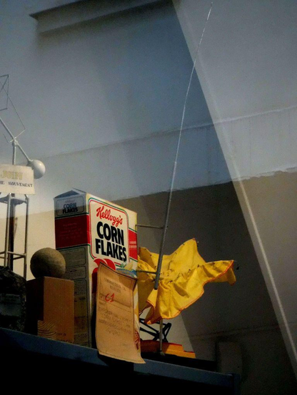 A Kellogg’s Corn Flakes box sits on a high shelf alongside assorted objects, including a stone on a wooden block, wire sculptures, a yellow fabric piece, and a long thin antenna rising awkwardly above the box.