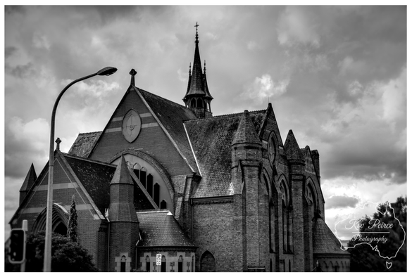 A striking black and white, low angle photograph of a historic church in Launceston, Tasmania.  The building is constructed of dark brick in a Gothic Revival style, featuring steep slate roofs, buttresses, and tall, narrow arched windows.  A small spire topped with a cross rises from the center of the roofline. The dramatic sky is filled with heavy, textured clouds, creating a strong contrast with the architecture.  A modern streetlamp and some dark foliage are visible on the left side, slightly framing the building.