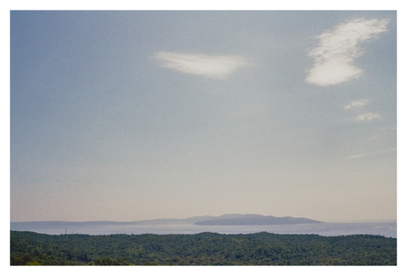 Color photograph of the Adriatic Sea with the island of Cres, taken from a viewpoint above the forests surrounding Kastav. The treetops of the green forest fill the lower part of the image. Above a narrow strip of sea and island elevation, a hazy sky with a few scattered clouds extends across most of the photograph.