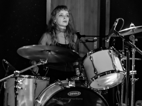 A female drummer performs on stage, seated behind a drum kit. The image is in black and white, showcasing her focused expression and long hair. The background features dark walls and stage equipment.