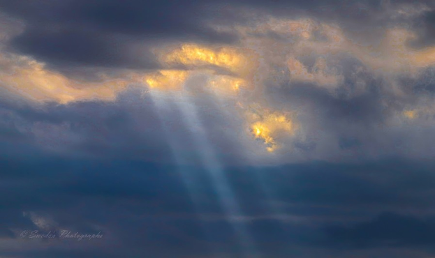 "The sky is a dramatic canvas of dark, brooding clouds—thick and layered, like a gathering of silent witnesses. From a break in the cloud cover, beams of sunlight burst through in radiant streaks, casting golden shafts across the gloom. These crepuscular rays fan out like divine brushstrokes, illuminating the edges of the clouds with a warm glow that contrasts sharply with the surrounding gray. The light feels directional, purposeful—like a celestial spotlight piercing the veil. The background is soft and diffuse, suggesting a wintry or overcast day, and the overall mood is reverent, cinematic, and quietly powerful. It’s a moment of atmospheric tension and grace, where shadow and light negotiate the sky." - Microsoft Copilot