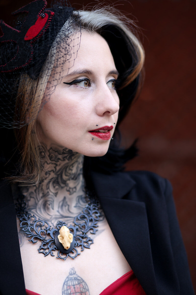 An alternative model's close portrait. She is wearing a black and red outfit. Her neck's black tattoo and necklace reinforce the baroque tone of the portrait. Around her red lips, we can see a canine bite piercing.

The blurry red-ocher tone background keeps the dominant tone of the portrait.