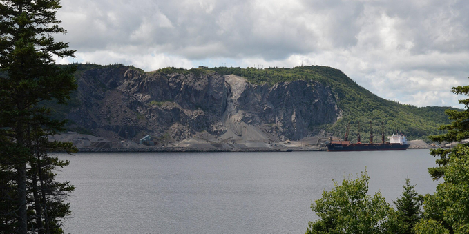 A photo of a strait with a mountain being quarried on the far shore. There is a ship in the strait. The sky is cloudy.