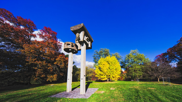 About 40% from the left are concrete pillars rising out of a pad with top-heavy structures on top,  this is in the middle of close-cut grass with reddish trees on the edge of the field to the left and a bright yellow one far away near the center with greenish trees behind that and something brown or red to the right