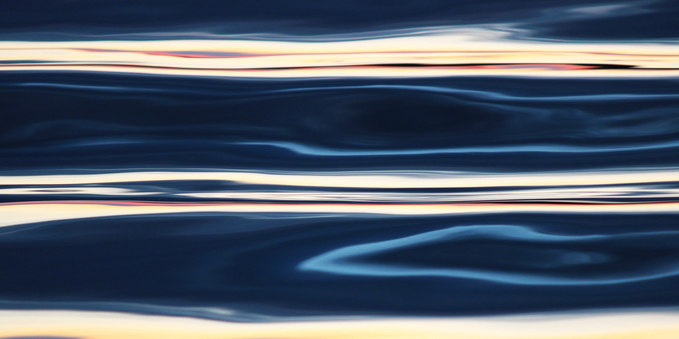 Photograph of sunset light reflected on water forming two bright horizontal stripes contrasting with dark and light blue stripes and swirls between them.