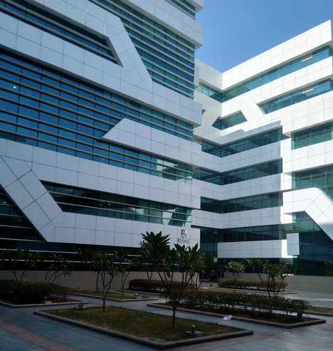 Modern office building with geometric white panels and glass windows under a clear blue sky. Foreground features landscaped areas with small trees.