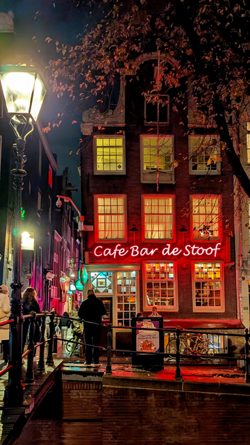Colour photo of a bar by a canal in Amsterdam, Netherlands. Image shows a brightly lit building with "Cafe Bar de Stood" in red neon lights.
Bar is on a canal bridge crossing, there's an old fashioned street lamp on the bridge.
Autumnal leaves show on a tree top of image and people are walking around the narrow side streets to the left.
Taken 19 November 2025.