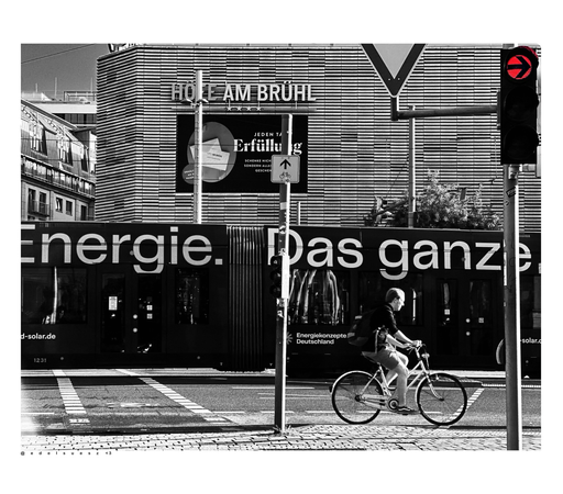 Aufnahme in schwarzweiß mit einem farbigen Akzent, Innenstadt Leipzig, eine rote Ampel leuchtet, seitlich ein vorbeifahrender Radfahrer, dahinter eine vorbeifahrende Straßenbahn mit großflächiger Werbung „Energie. Das ganze…“ im Hintergrund die Fassade eines Einkaufszentrums mit der Aufschrift „Höfe am Brühl“ 
