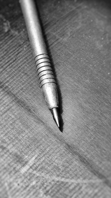 Detailaufnahme in Schwarz-Weiß vom vorderen Teil einer Reißnadel aus Metall, bestehend aus Schaft und Spitze. Der Schaft der Reißnadel verläuft von links oben leicht schräg nach unten bis zur Bildmitte, wo er in der Spitze endet. 