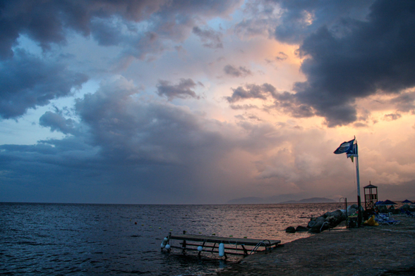 The image shows a coastal scene at sunset, featuring a darkening sky filled with clouds, a calm sea, and a small dock extending into the water. A flag is visible on the shore, with rocks and lounge chairs in the foreground.