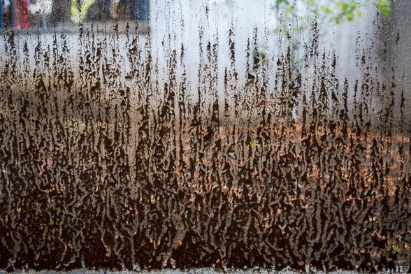 In the foreground of the photo is a glass pane covered in splashes of mud that form intricate patterns. Behind the muddy glass, a background of what seems to be a house is vaguely visible.