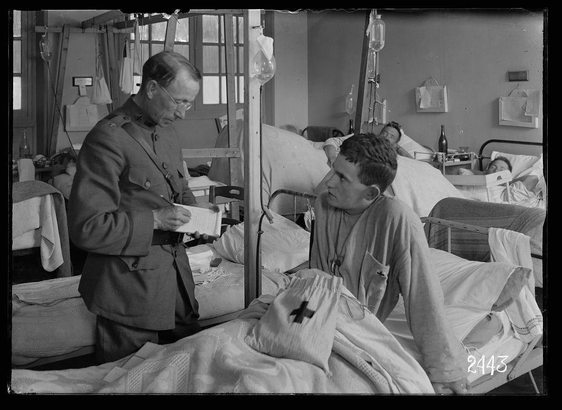 The image depicts a black and white scene within what appears to be an old military hospital ward. A figure dressed in a uniform with insignia indicative of the Army Medical Corps is standing beside a bed, attending to another individual lying down who seems to have been injured or ill; he wears clothing marked by a red cross indicating that they are likely part of the Red Cross medical service for soldiers and civilians alike during wartime periods. The surrounding area shows multiple beds occupied with other patients receiving treatment through intravenous lines as suggested by hanging saline bags, along with personal items like bottles and newspapers scattered around their quarters.

In terms of historical context provided in external resources, this photograph likely dates back to World War I or II given the style of uniforms and hospital equipment. It was taken at a military medical facility in Neuilly during that era when Rev Father Wareing served as a chaplain for ARC (American Red Cross), which operated hospitals on battlefields throughout various global conflicts including WWI, WWII, Korea, Vietnam, etc.

The presence of bottles suggests these could be medicinal or disinfectant beverages typically used to clean wounds and aid in healing. The overall setting is indicative not only of the medical care available but also reflects wartime conditions where soldiers might have been treated alongside civilians if resou [...]