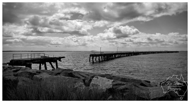 A black and white photograph of the historic, partially dilapidated Tanker Jetty in Esperance, Western Australia.  The remains of the jetty stretch out into the ocean under a dramatic, cloudy sky. In the foreground, large rocks and some coastal grass frame the image.