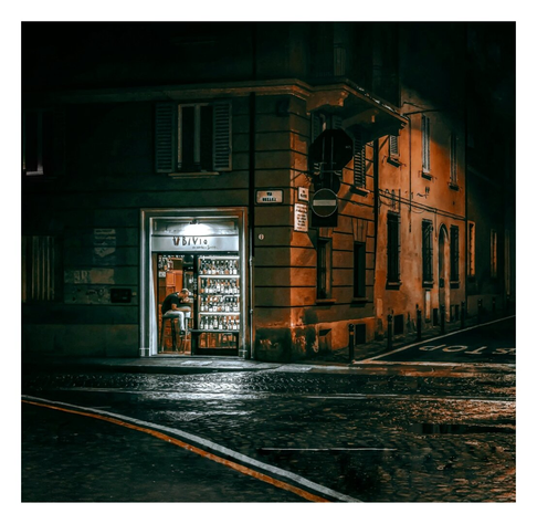A photo of a dimly lit, narrow street corner at night. A small, warmly lit wine shop with a "Bivio" (the crossroads) sign is centered, its display window filled with various winebottles. The building is aged, with weathered brown/orange and grey walls, shutters, and a streetlight casting orangey light. The wet cobblestone street reflects the light. The background is shadowy, with faintly visible neighboring buildings.