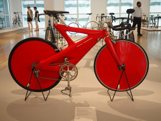 Ein rotes Karbon-Rennrad von 1989 in einer Fahrrad-Ausstellung. Im Hintergrund, gr0ße Fenster, weitere Fahrräder und Pblikum.