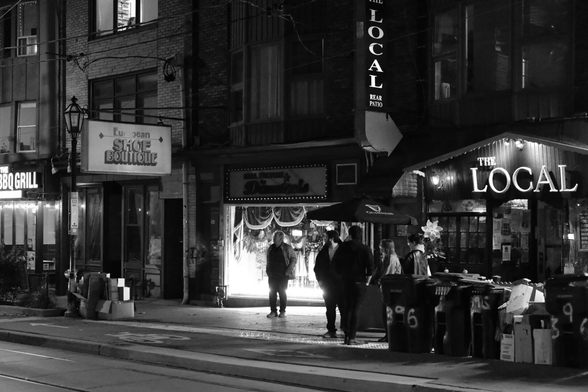 Night outside The Local pub. People mill around on the street outside, maybe smoking. Next door, there’s a women’s wear store with a brightly lit window and a person is framed in the lights. Along the street, the recycling bins are out awaiting pickup in the morning.