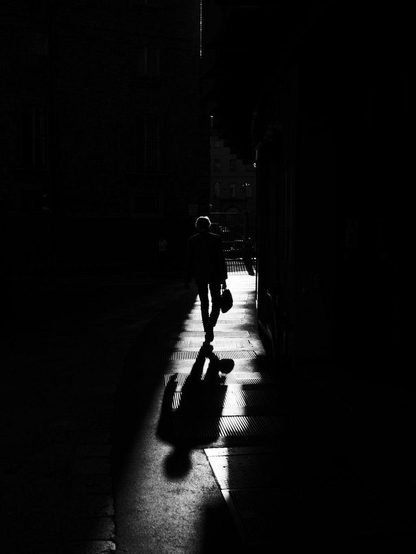 A black and white photograph of a man with a suitcase walking away from us in a sliver of harsh sunlight among shadows projected by tall buildings.