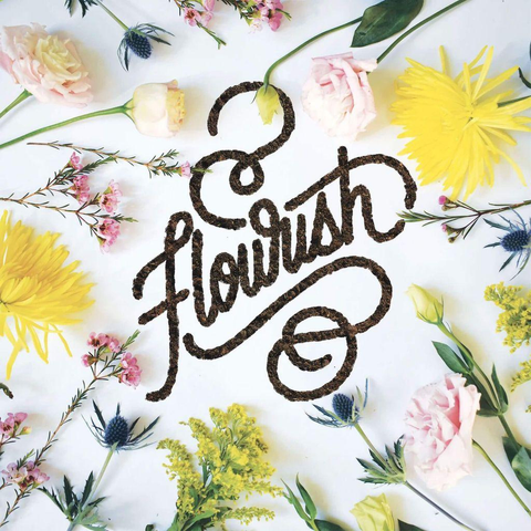 The word "Flourish" crafted from coffee grounds is surrounded by vibrant flowers of various colors and types on a white background.