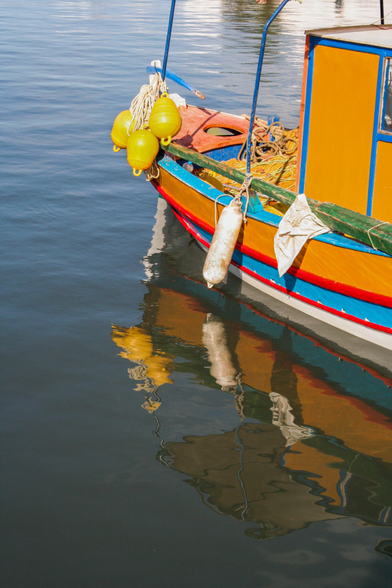 A colorful boat with yellow buoys and fishing gear is moored. The boat's reflection is visible in the still water.