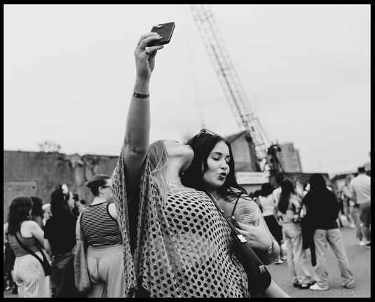Black and white photograph of a woman holding a smartphone aloft to take a selfie with another woman at a crowd gathering, with industrial dock cranes and buildings visible in the background, captioned "Two women taking a selfie at the Cork Pride party on Kennedy Quay last August".