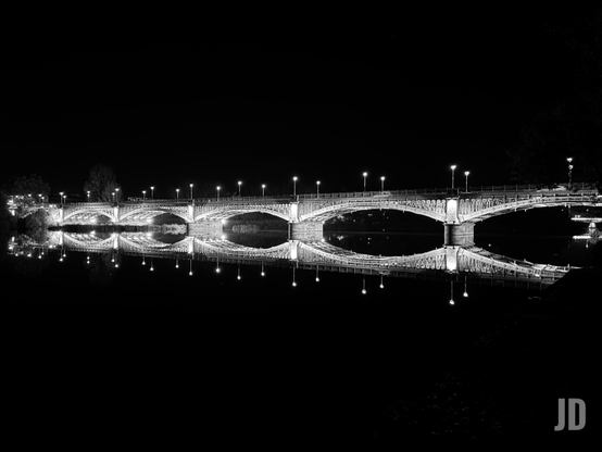 Una fotografía nocturna en blanco y negro de un puente arqueado sobre el agua. El puente presenta múltiples arcos semicirculares y está brillantemente iluminado por una fila de farolas encendidas que se extienden a lo largo de su parte superior. El agua, muy tranquila, crea un reflejo casi perfecto del puente y sus luces, lo que resulta en una composición simétrica que forma una figura similar a un símbolo de infinito o un ocho horizontal. El cielo y el agua circundante son completamente oscuros, resaltando el brillo del puente y sus reflejos.