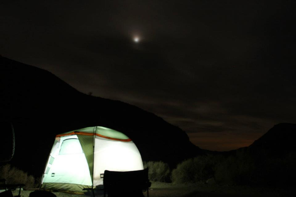 In the nighttime darkness, an internally illuminated tent sits below the moon high up in the clouds.