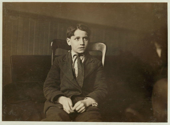 The image is a black and white photograph of a young boy sitting in an armchair. He appears to be around 12 years old, based on the provided information about Joseph Buonasari's age during that time period.
Joseph is wearing a suit with a tie, which suggests he may have been dressed up for a special occasion or perhaps this was taken as part of his school records. His expression seems contemplative and somewhat serious.
The background appears to be indoors, possibly in an office setting given the presence of what looks like blinds on a window behind him. There is another person partially visible in the frame, but only their arm can be seen resting on a surface next to Joseph's chair.
Additional context about this image comes from documents mentioning Joseph Buonasari's life details at that time: he worked various jobs such as beans and apples factory work for 50 cents per day. His parents also had challenging working conditions with extended hours. This photograph was taken in Buffalo, New York (State) on January 11th.
The image has a timestamp of '207_ThirteenthStreet_UniversityOfChicago', indicating it may be part of an archival collection or associated research project from the University of Chicago.