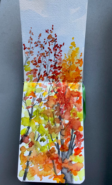 A small, narrow sketchbook, vertically shown, with autumn trees painted in watercolor across the two pages. Reds, oranges, yellows and greens. 