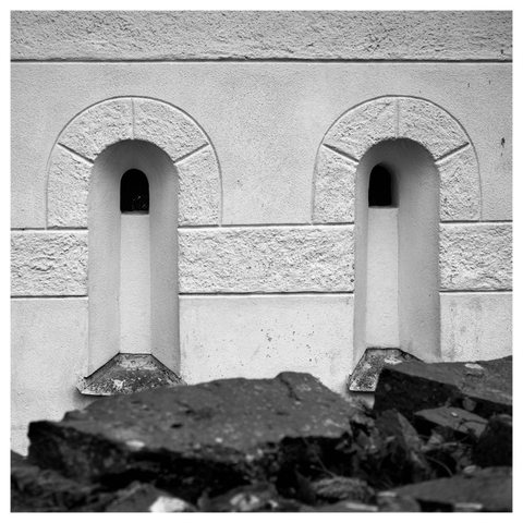 En fyrkantig svartvit bild på två smala vertikala fönster i en gammal, vit stenbyggnad. Ovansidan är rundad och nedre sidan har utstickande fönsterbleck. I förgrunden en mörk stenmur.