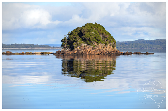 A tranquil, wide shot photograph of a small, dome shaped rocky island covered in dense green scrub and trees, resting in the smooth, mirror like blue waters of Macquarie Harbour.

The island's reflection is perfectly cast on the surface of the water, and the distant shore, covered in forest, sits under a bright blue sky with scattered white clouds. Photo by Kev Peirce.