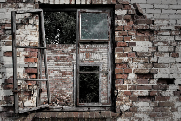 Verfallene Backsteinmauer mit zwei rechteckigen Fensterrahmen ohne Glas, dahinter Bäume sichtbar.