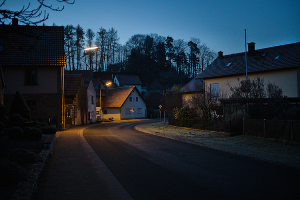 Straße führt von unten Mitte nach rechts durchs Bild. An beiden Seiten Häuser. Dorflampnen Leuchten. Blaue Std. Frost auf der Wiese und Dächern.