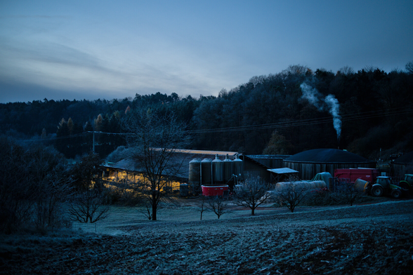 Ein beleuchteter Stall in der Ferne. Große Behälter aus Metall. Am Anfang. Rechts die Biogas Behätnisse mit gebogenen Dach aus Gummi. Ballen Trecker und Ahnhänger rechts an der Seite. In Vordergrund ein Feld. Im Hintergrund Wald. Eine Strohmleitung übern Stall. Rauch vom Motor bei den Gas Behältern. Im Bild alle smit Frost bedeckt.
