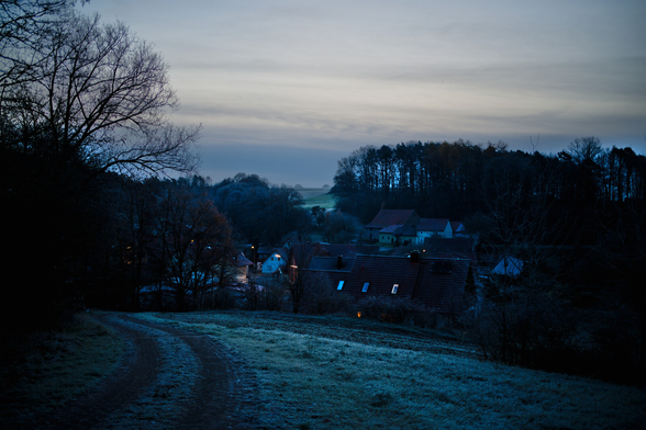 Ein Dorf im Tal. Von oben Nach unten Fotografiert. Dorflampen leuchtne. Ausenrum Waldstücke und Wiesenstücke. Frost überall. Der Himmel mit Wolkenstreifen.